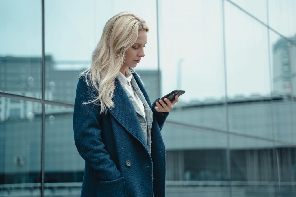 pexels-photo-5717224-5717224 Professional woman in blue coat using smartphone outside office building.